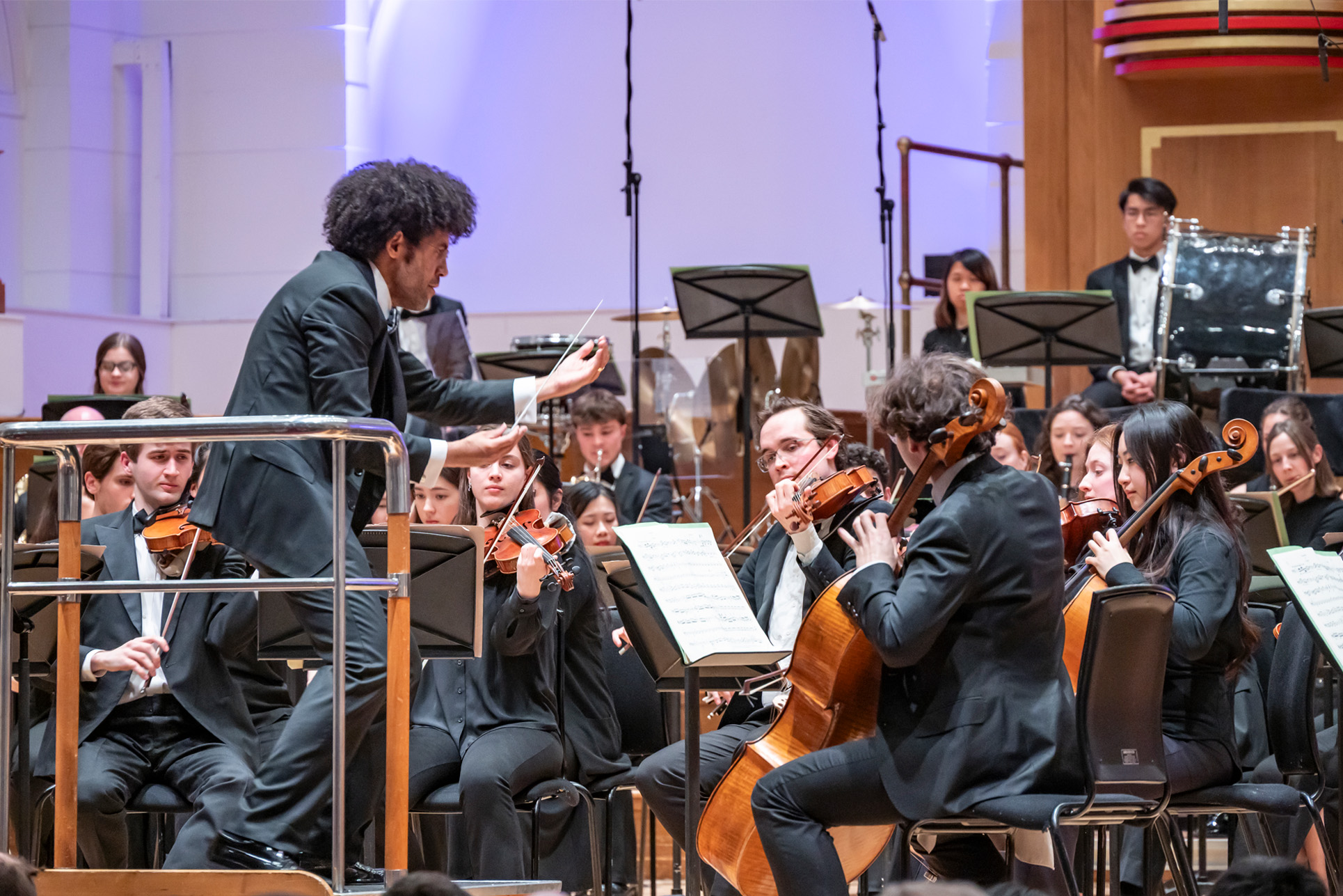 A man wearing a black smart suit, conducting an orchestra, with the cello section closest to him, all wearing smart attire.